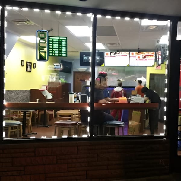 Exterior view of AlBasha Subs at night, showing customers and staff inside through the window.