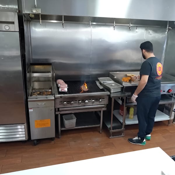 AlBasha Subs chef grilling meat and chicken on a large stainless steel grill in a commercial kitchen.