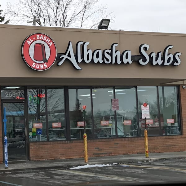 Exterior of AlBasha Subs restaurant with its sign, large windows, and brick facade.