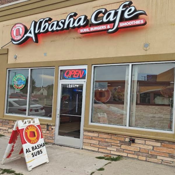 Exterior of AlBasha Cafe with a neon sign, an "OPEN" sign, and an "Al Basha Subs" sandwich board.