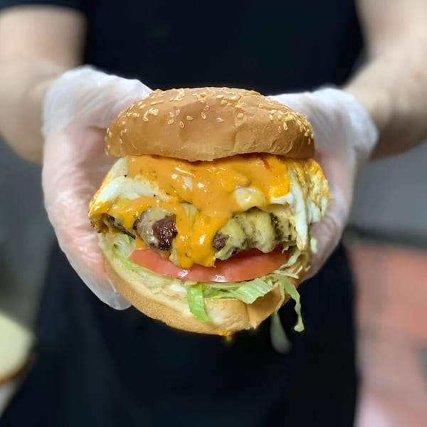 Gloved hands hold a juicy AlBasha burger with melted cheese, fried egg, lettuce, and tomato.