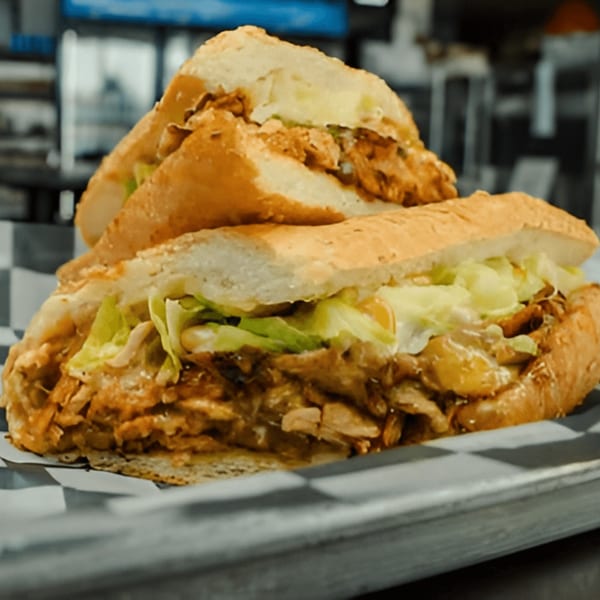 Two AlBasha Subs, filled with meat, cheese, and lettuce, on a checkered paper-lined tray.