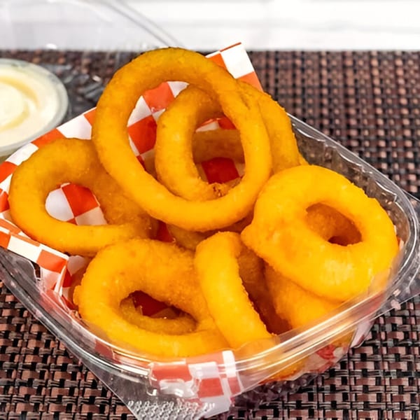 Golden-brown onion rings in a clear plastic container with red and white checkered paper, next to a dipping sauce.