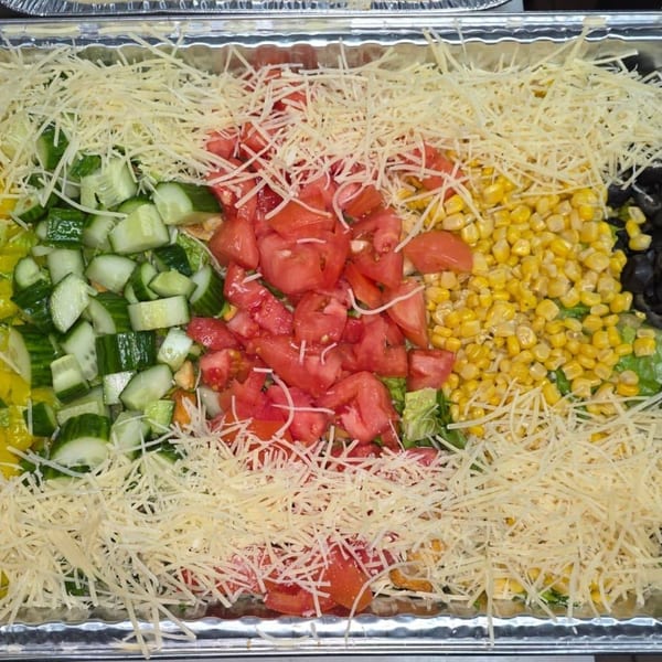 A catering tray filled with salad, topped with shredded cheese, cucumbers, tomatoes, and corn.